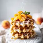 Tall golden Belgian waffle stacked with glossy macerated peach slices dripping with melted brown butter, dusted with powdered sugar, and garnished with a fresh green mint sprig on a bright morning plate setting.