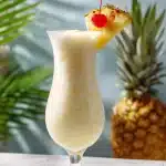 A tall frosty pina colada in a hurricane glass garnished with a fresh pineapple wedge and a cherry, against a sunny tropical backdrop.