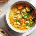 A warmly lit white bowl of creamy golden prebiotic vegetable soup, filled with visible leek rings, white beans, carrot half-moons, and Jerusalem artichoke cubes, garnished with fresh parsley and served with a side of crusty bread on a bright marble surface.