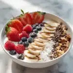 Vibrant probiotic yogurt breakfast bowl with bananas, blueberries, granola, almond butter drizzle, and chia seeds on white marble.