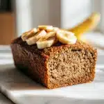 A thick slice of moist protein banana bread with a dense, tender crumb and golden-brown crust, topped with fresh banana slices and a light dusting of cinnamon, resting on a white plate in bright, natural window light.
