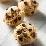 A bowl of creamy no-bake protein dessert cookie dough with dark chocolate chips on a light marble surface, lit by natural window light.