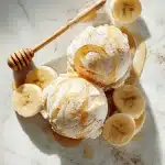A scoop of creamy protein ice cream drizzled with golden honey, surrounded by fresh banana slices and a dusting of cocoa on a bright marble surface.
