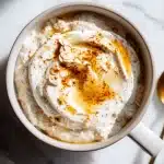 A white bowl of creamy protein overnight oats topped with a dollop of Greek yogurt, a swirl of cinnamon, and a drizzle of golden maple syrup on a bright white marble surface.
