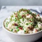 A creamy resistant starch potato salad with fresh dill, green peas, crispy bacon, and a dollop of Greek yogurt, on a white marble surface.