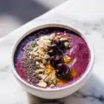 A vibrant deep purple sleep recovery smoothie bowl topped with oats, pumpkin seeds, chia seeds, whole tart cherries, and honey drizzle on a bright marble surface.