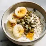 Warm bowl of creamy oatmeal with banana slices, honey drizzle, pumpkin seeds, and flaxseed on marble surface, sleep supporting oatmeal.