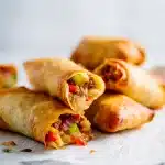 Close-up of crispy steak egg rolls with golden-brown wrappers, oozing provolone cheese, and colorful peppers on a white marble background.