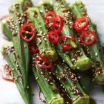 Fresh steamed okra with garlic-chili sauce, red chili slices, sesame seeds, showcasing steamed okra recipes without slime on a bright surface.
