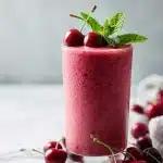 A glass of creamy tart cherry sleep smoothie topped with fresh cherries and mint leaves on a marble counter, with a soft, bright setting.
