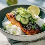 Glossy teriyaki salmon fillet over fluffy rice, topped with quick-pickled cucumber coins, scallions, sesame seeds, and a lime wedge.