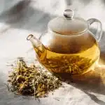 A warm glass teapot of golden valerian root tea with dried herbs and honey on bright linen, showcasing a calming valerian root tea recipe.