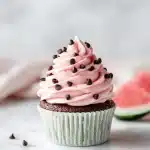 A close-up of a pink buttercream-frosted cupcake decorated with chocolate chip seeds and a small watermelon slice garnish, sitting on a white plate with a bright, airy summer backdrop.
