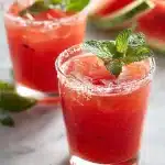 A tall glass of watermelon lemonade with ice, fresh mint leaves, and a salted rim, garnished with a watermelon wedge, sitting on a wooden table outdoors in sunlight.