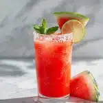 Fresh watermelon margarita in salt-rimmed glass with lime wheel, watermelon wedge, mint sprig, on bright marble surface with tequila bottle.