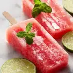 Two fresh watermelon popsicles on wooden sticks, garnished with lime slices and mint leaves, showing icy pink crystals. Close-up.
