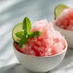 Fluffy pink watermelon shaved ice in a white bowl, topped with fresh green mint leaves and a lime wedge on a wooden table.