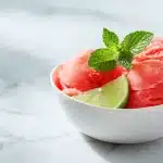 Three scoops of watermelon sorbet in a ceramic bowl garnished with mint and lime