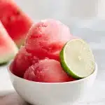 A scoop of bright pink watermelon sorbet with a lime wedge garnish, served in a minimalist white bowl on a light marble surface, refreshing summer dessert.