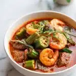 Steaming bowl of west african okra stew recipe with sliced green okra, pink shrimp, beef chunks, red palm oil broth, and herb garnish.