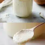 A clear glass jar filled with a creamy, bubbly Amish Sourdough Starter, placed on a rustic wooden table. Beside the jar are scattered ingredients including a small bowl of flour and a glass of milk, highlighting the sweet, yeasty, living texture of the friendship bread base.