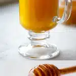 A steaming glass mug of golden honey cinnamon ginger tea on a wooden table, surrounded by scattered cinnamon sticks, fresh ginger root slices, and a honey dipper, with soft natural morning light.