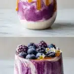 A tall glass of thick Greek yogurt fruit smoothie with vibrant purple mixed berry layers, bright white yogurt ribbons swirling through, and a glossy honey drizzle on top, sitting on a clean white marble surface with scattered fresh berries.
