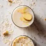A clear glass of creamy, milky-white rice water with thin slices of fresh ginger floating inside, placed on a bright white marble countertop. Scattered raw white rice grains surround the base of the glass, with soft natural light highlighting the soothing drink.