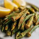 A light marble platter holds golden-brown, crispy oven-roasted okra pods seasoned with smoked paprika, next to bright yellow lemon wedges. This roasted okra recipe makes a perfect, slimy-free side dish.