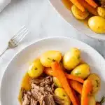 A fork-tender slow cooker beef joint with glossy brown gravy, surrounded by vibrant orange carrot chunks and golden Yukon Gold potatoes, styled on a bright white marble surface with fresh herb garnish.