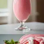 A tall glass filled with a thick, vibrant pink strawberry smoothie, garnished with a whole fresh strawberry on the rim and a swirl of white yogurt on top, sitting on a marble counter with scattered strawberries nearby.